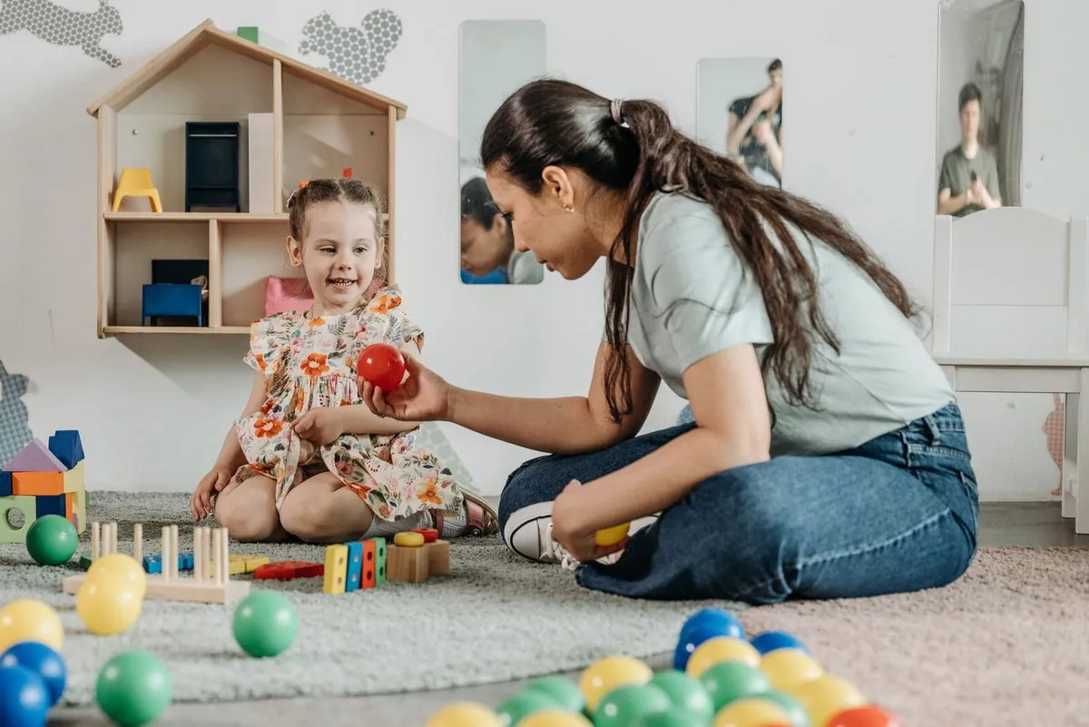 Terapeuta sentada no tapete brincando com uma menina sorridente, cercadas de bolinhas coloridas e brinquedos de madeira