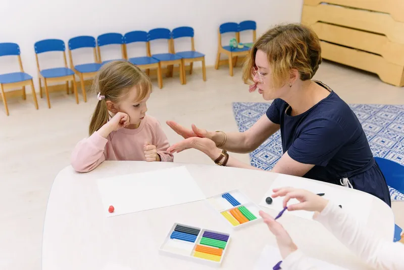 Terapeuta modelando massinha colorida em uma mesa junto com uma menina atenta à atividade